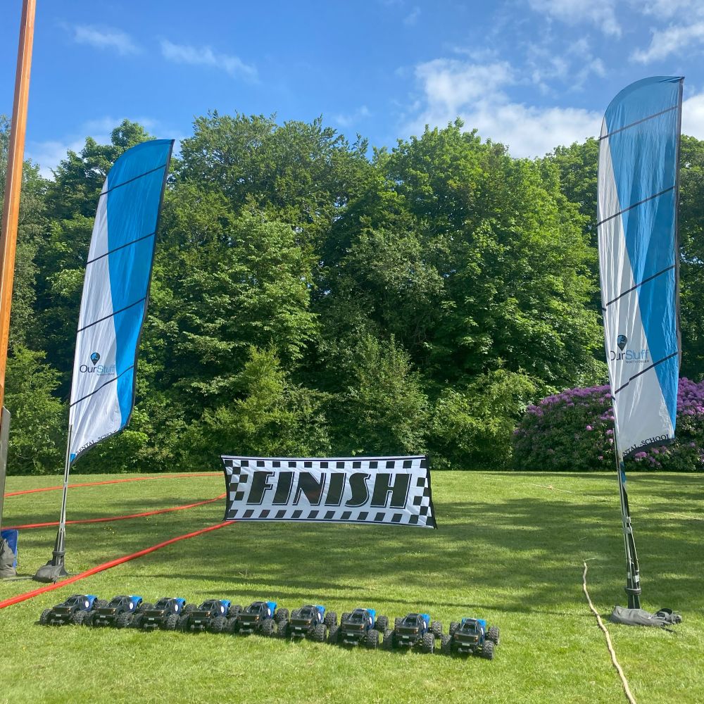 En række fjernstyrede biler på en græsplæne med en mållinje i baggrunden. At køre med Radio Control Cars er en sjov oplevelse for alle. I inddeles på hold, så I kan få en fornemmelse af konkurrence, samarbejde og har det sjovt sammen.