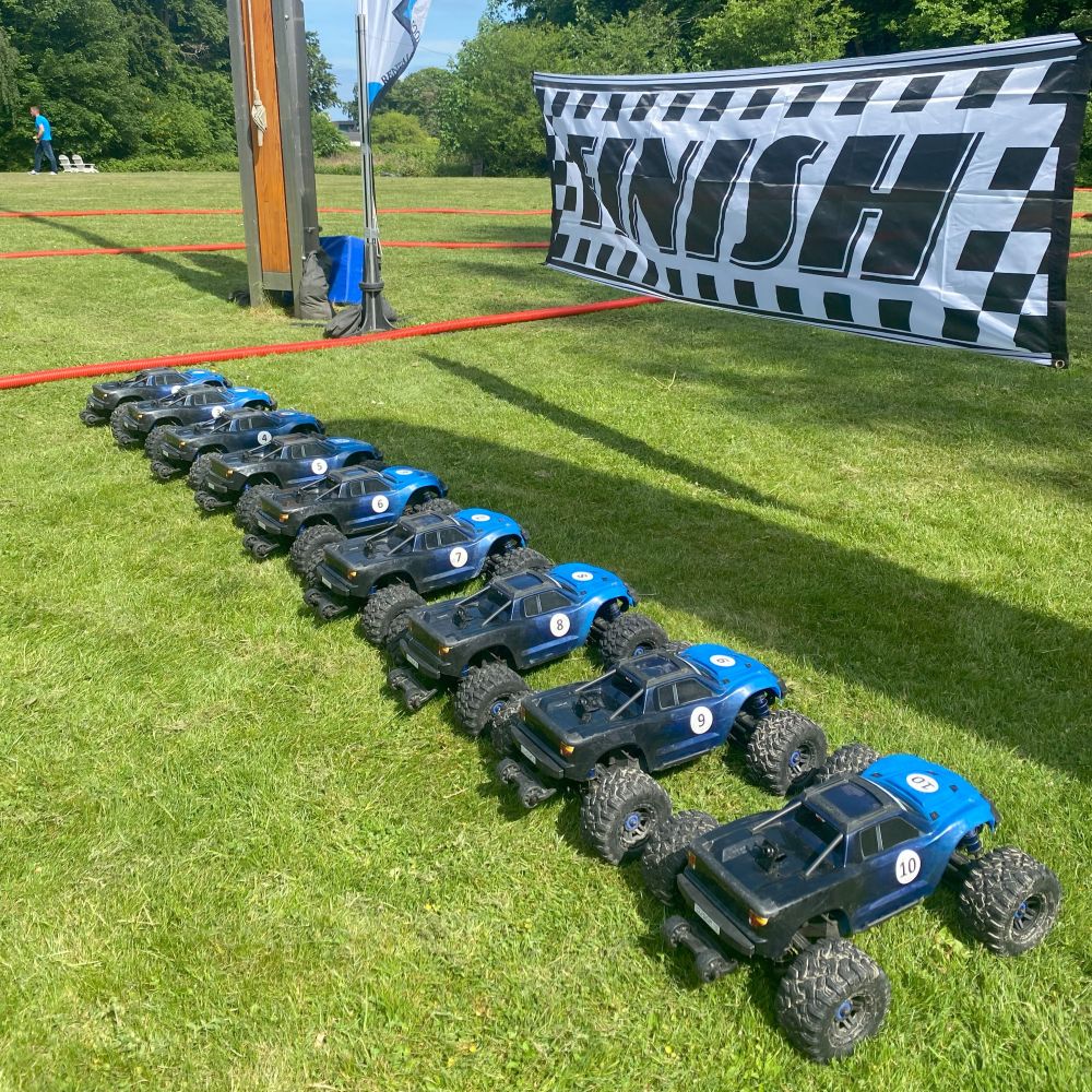 En række fjernstyrede biler på en græsplæne med en mållinje i baggrunden. At køre med Radio Control Cars er en sjov oplevelse for alle. I inddeles på hold, så I kan få en fornemmelse af konkurrence, samarbejde og har det sjovt sammen.