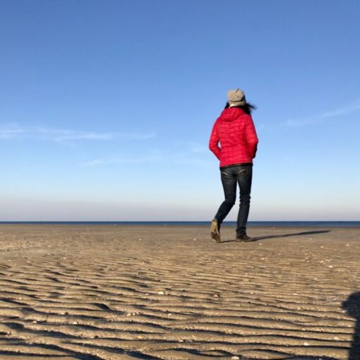 En kvinde i rød jakke og grå hue går på stranden på Rømø