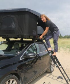 Ung kvinde står på stigen til et tagtelt monteret på en sort bil, der står parkeret på stranden