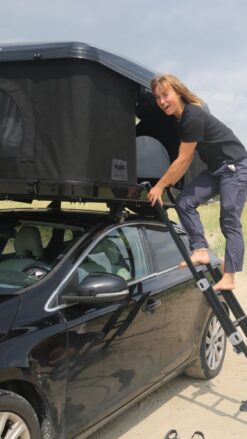 Ung kvinde står på stigen til et tagtelt monteret på en sort bil, der står parkeret på stranden