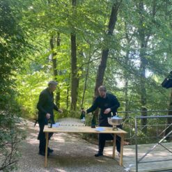 Midt på en skovsti er et bord sat op. To mænd er i gang med at hælde champagne op til gæster ved et firmaarrangement.