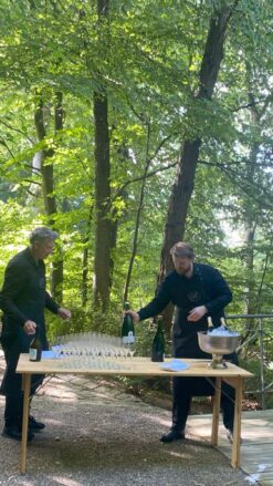 Midt på en skovsti er et bord sat op. To mænd er i gang med at hælde champagne op til gæster ved et firmaarrangement.