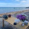 Et solidt træbord er sat op på stranden med drikkevarer og frisk afskårne blomster. Havet og en badebro ses i baggrunden. Afventer gæster til et firmaevent.