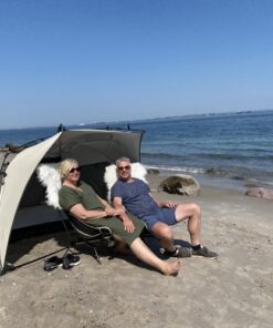 Et midaldrende par sidder på stranden i behagelige stole med et lætelt. Der er blå himmel