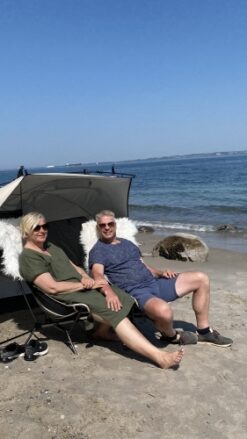 Et midaldrende par sidder på stranden i behagelige stole med et lætelt. Der er blå himmel