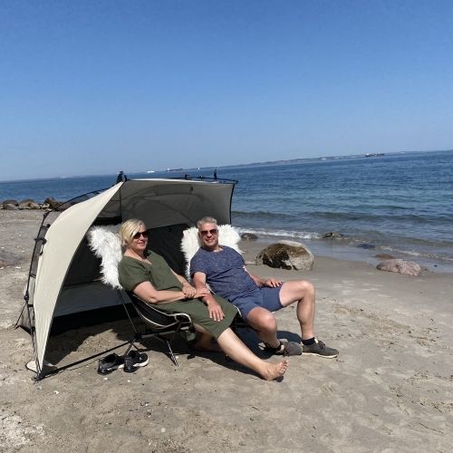 Et midaldrende par sidder på stranden i behagelige stole med et lætelt. Der er blå himmel