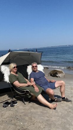 Et par sidder i stole fra Nordisk beklædt med blød "pels" under et læsejl lige ved vandkanten.