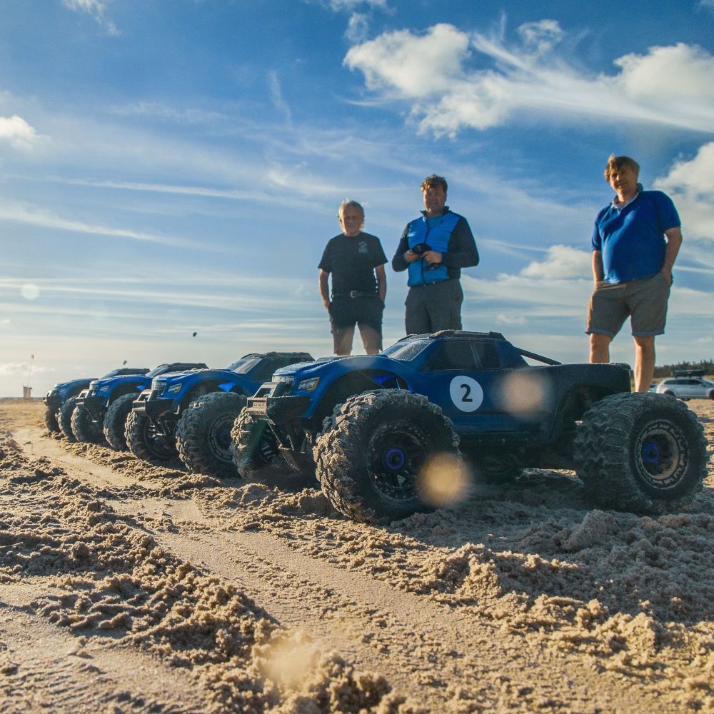 Nærbillede af en række fjernstyrede biler på en strand med blå himmel og sol i baggrunden. At køre med Radio Control Cars er en sjov oplevelse for alle. I inddeles på hold, så I kan få en fornemmelse af konkurrence, samarbejde og har det sjovt sammen.