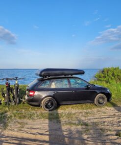 En sort bil med et sort tagtelt monteret og 2 cykler bagpå står parkeret med udsigt til havet