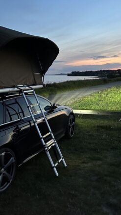 En sort bil med opslået tagtelt og stige parkeret med lygterne tændt ved solnedgangstid