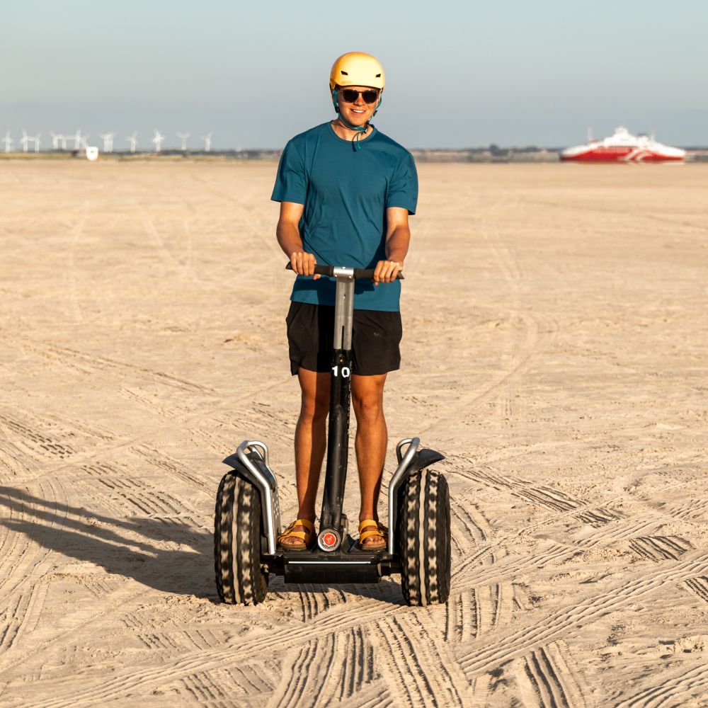 En ung mand kører på Segway på en strand, hvor solen skinner. Book jeres næste event med os og oplev, hvordan et Segway Event kan løfte teamets samarbejde til nye højder!