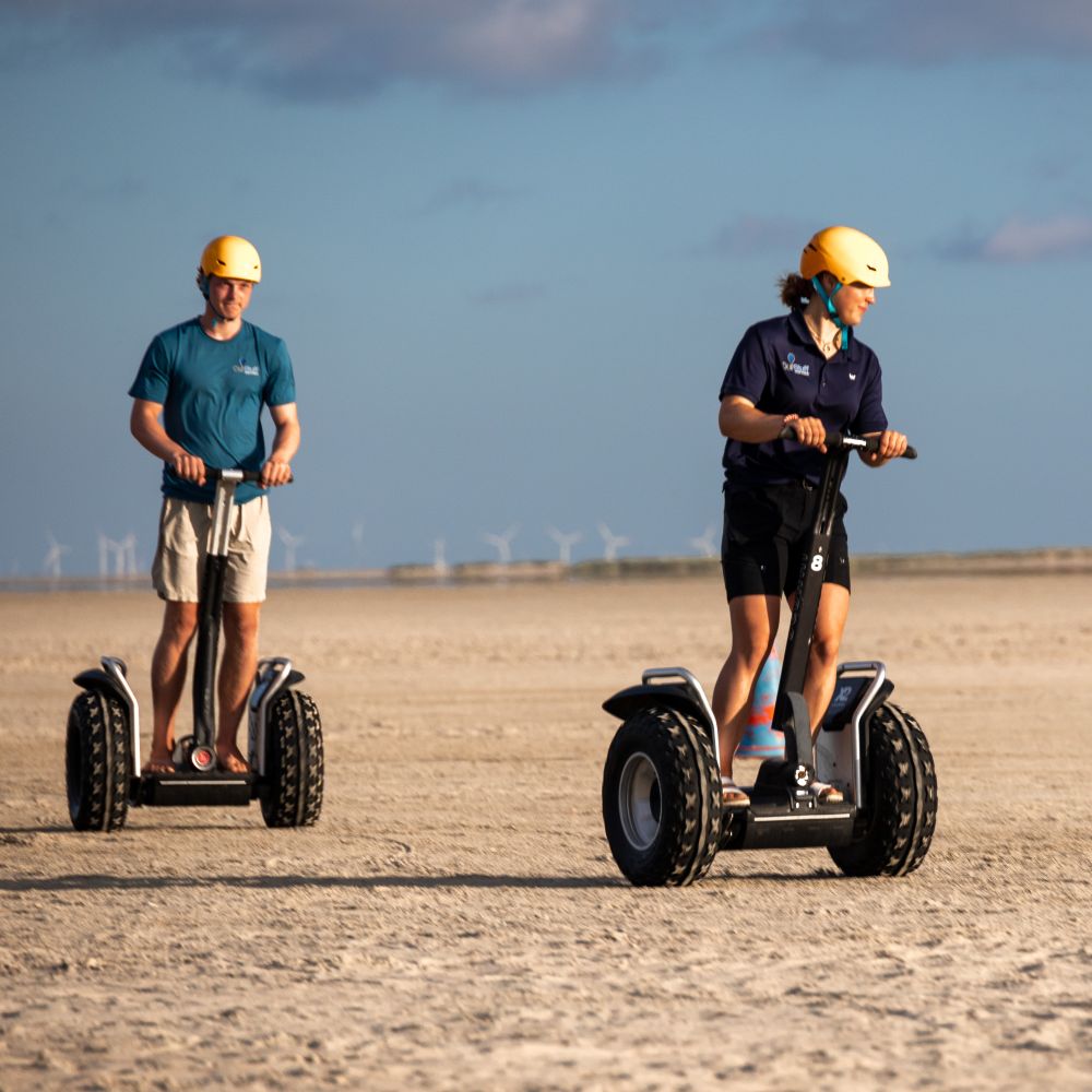 En ung kvinde og en ung mand kører på Segway på en strand, hvor solen skinner. Book jeres næste event med os og oplev, hvordan et Segway Event kan løfte teamets samarbejde til nye højder!