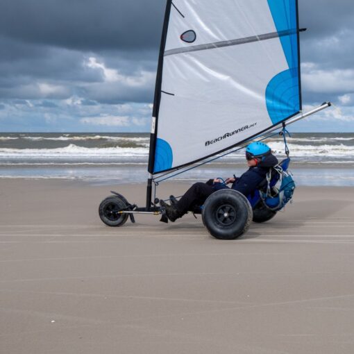En ung mand styrer sin strandsurfer hen over en flad strand. Der er grå skyer på himlen.