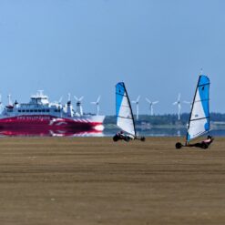 1 dobbelt og 1 single strandsurfer med hvidt og blåt sejl kører på en bred flad strand på Rømø- I baggrunden ses færgen til Sylt.
