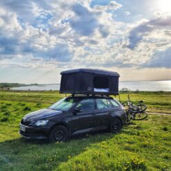 En sort bil med et sort tagtelt står på en græsplæne med kystlinje i baggrunden