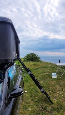 Et sort tagtelt er monteret på toppen af en sort bil. En stige fører til jorden. I baggrunden fisker en mand