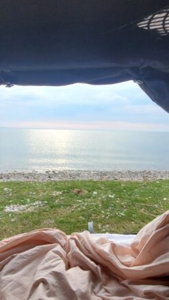 Udsigt over grøn græsplane og blankt vand fra et tagtelt, hvor der er redt op med dyner.